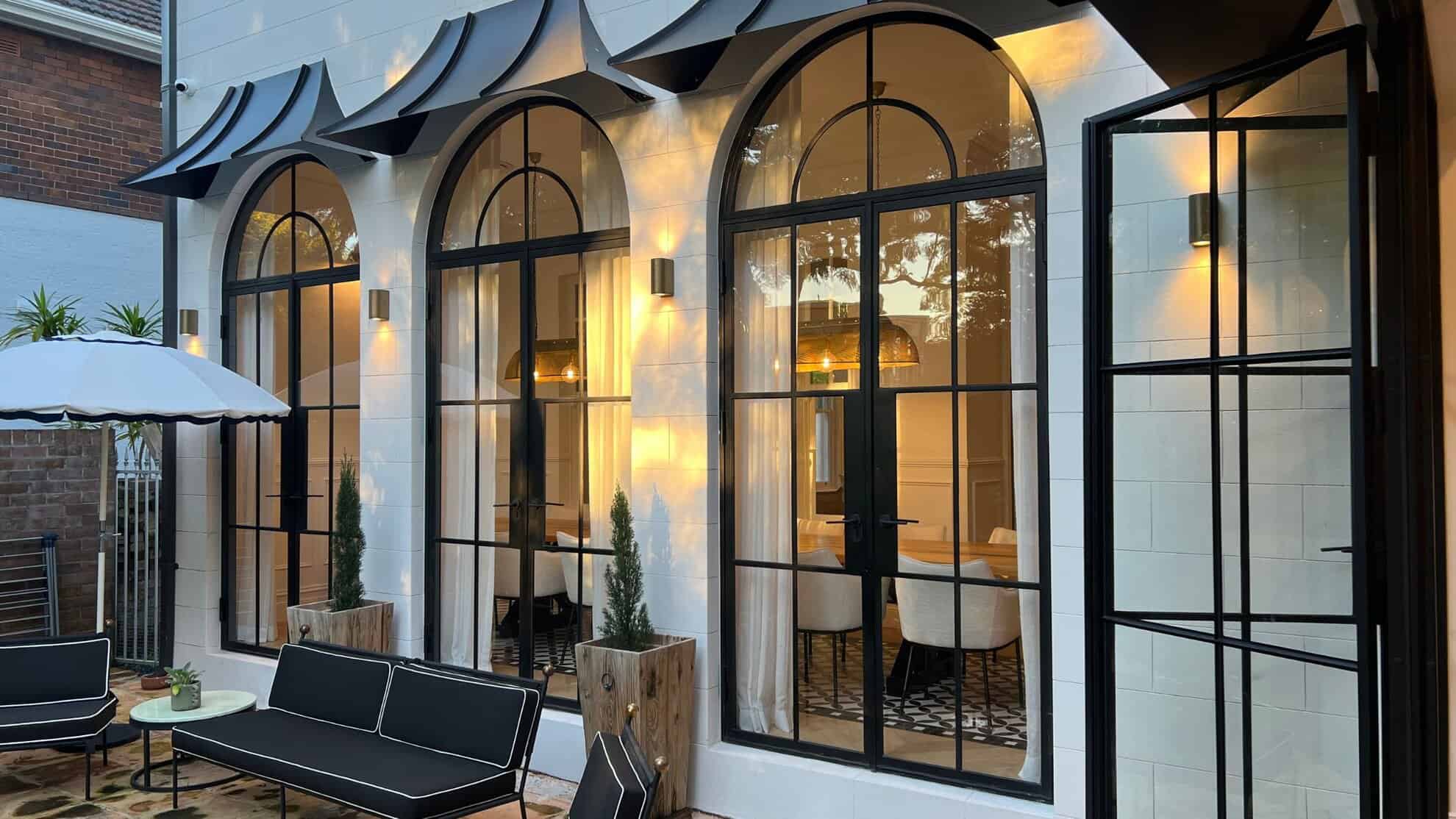 Lit stone facade with three arched black-framed doors and outdoor seating at dusk